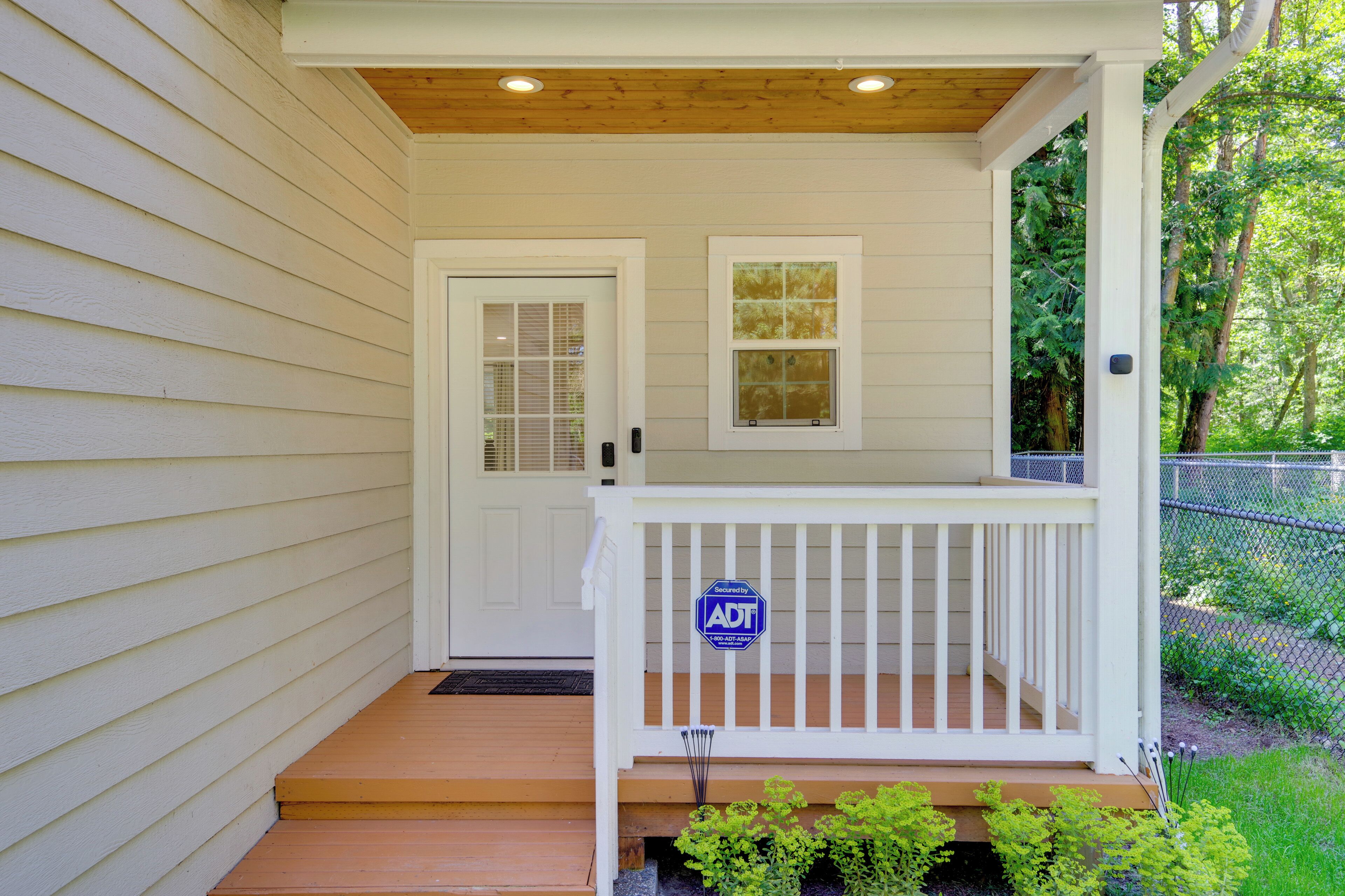 Une maison à l’extérieur blanc, avec un porche à l’avant doté d’une balustrade blanche, une porte avec une fenêtre et un panneau indiquant la présence d’un système de sécurité.