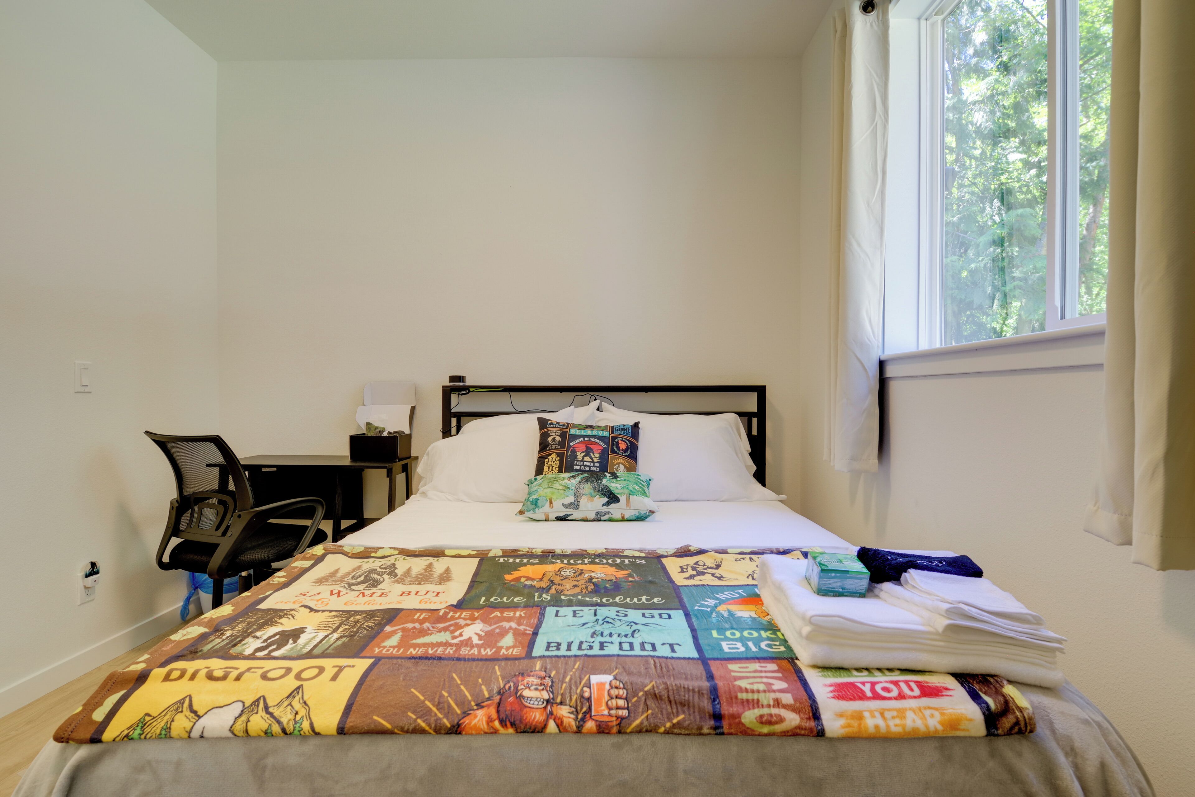 Una cama bien tendida con un colcha colorida, una silla, un escritorio y una ventana con cortinas.