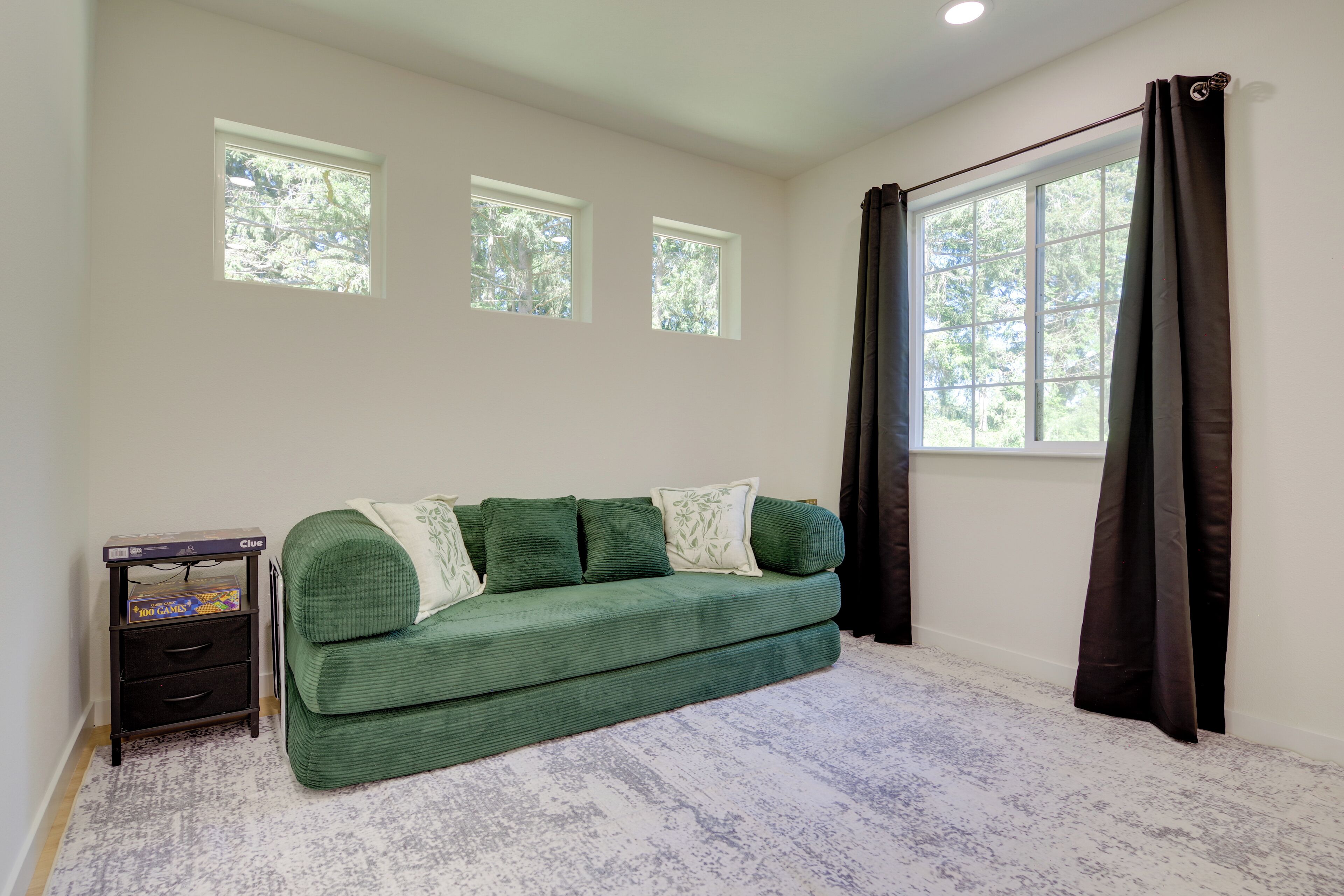 A room with a green sofa, a small side table with a board game, three windows, and a large window with curtains.