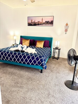 A hotel room with a large bed, a cityscape painting, a decorative wall hanging, a standing fan, and a small table with a lamp. - 2-bedroom apartment in fabulous Houston with cool AC (Houston)