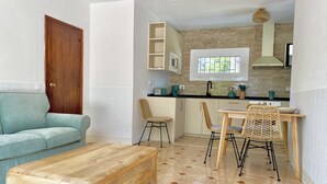 Un espacio habitable funcional con un banco de madera, un sofá color turquesa, una mesa de comedor con sillas de mimbre, una zona de cocina con ventanal y una puerta de madera.