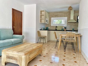 A compact living space with a wooden bench, a teal sofa, a dining table with wicker chairs, a kitchen area with a window, and a wooden door. - New Valparaiso house with private pool 8 people (Cunit)