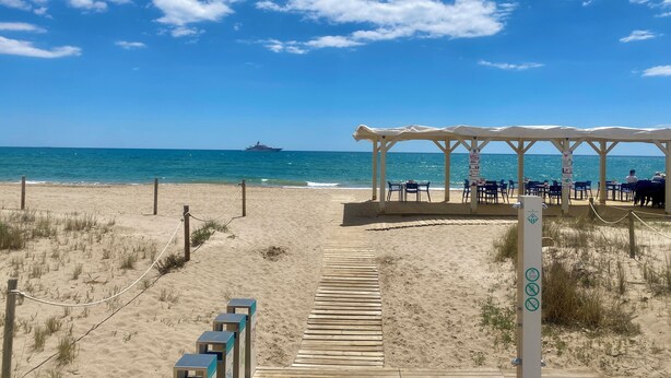 Una playa con un paseo de madera, un área de descanso techada y un muelle con un barco a lo lejos.