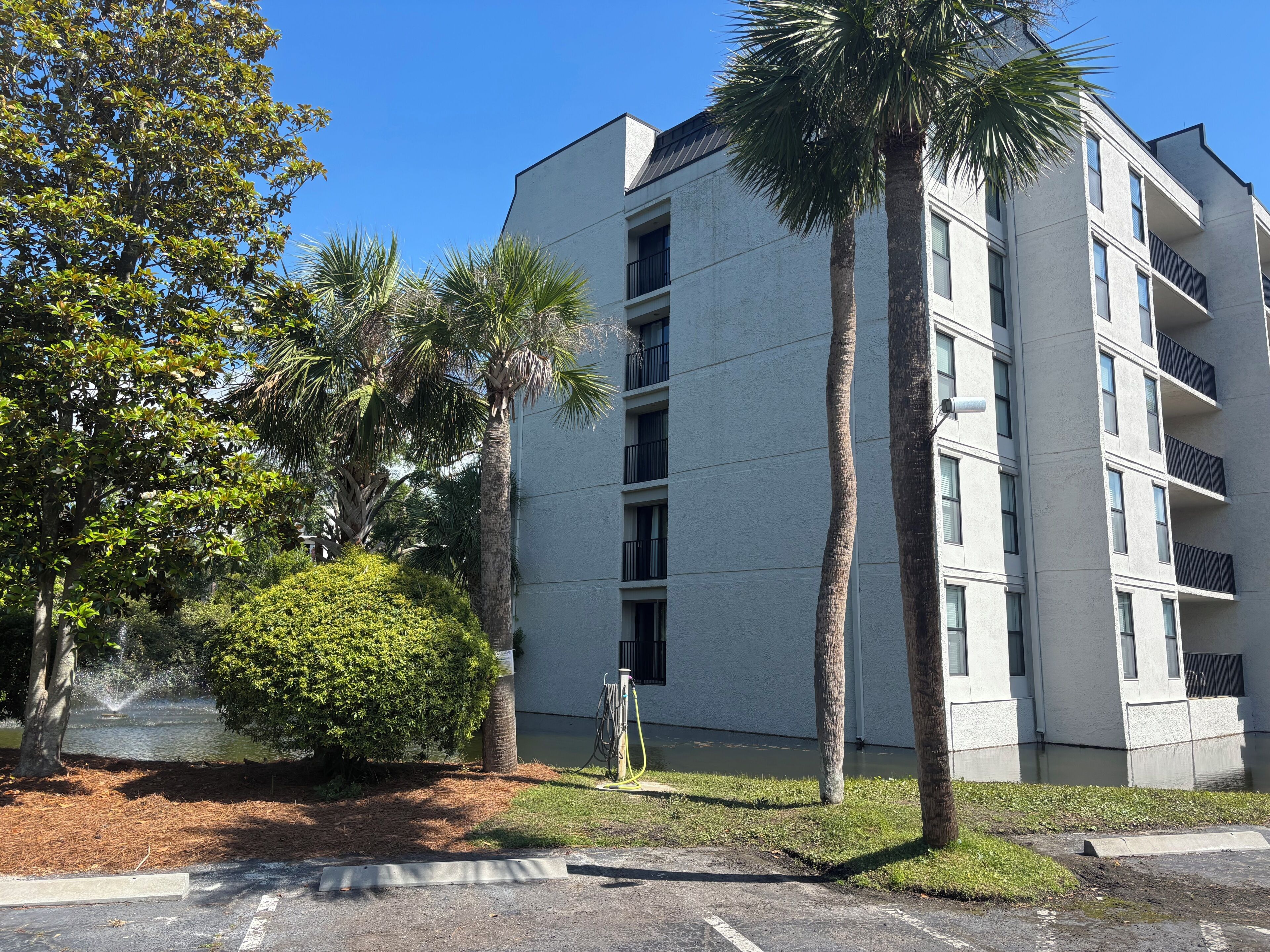 A multi-story building with balconies, surrounded by palm trees and a small water feature.