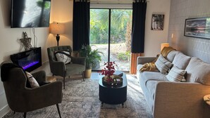 Ein Wohnzimmer mit Flachbildfernseher, einer Couch, einem Sessel, einem Couchtisch und Blick auf einen Garten durch eine Glastür.
