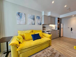 En moderne stue med en gul sofa, et lille bord og et køkken i baggrunden.