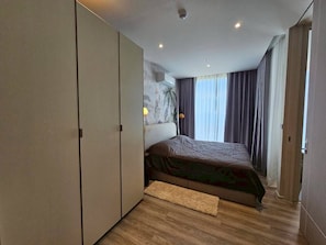 A bedroom with a bed, a wardrobe, a headboard with a mural, and a window with curtains. - Luxury 1BR Plus at Veha Hua Hin | Panoramic Sea View & Near Beach (Tambon Nong Kae)