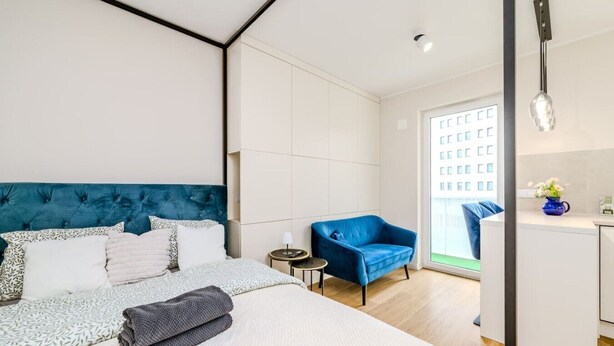 A modern hotel room with a large bed, a blue armchair, a small side table, and a view of a building through the window. - Wrocław Gaj Studio - Balcony - by Rentujemy (Wrocław)