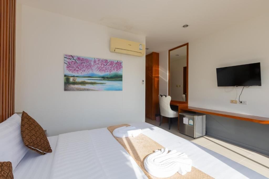 A hotel room with a bed, a painting, a desk, a chair, a TV, and an air conditioning unit.