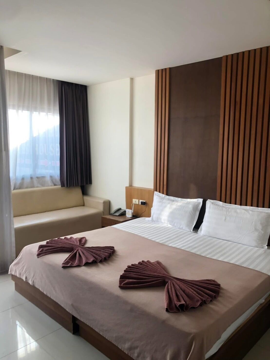 A hotel room with a bed, a bedside table, a lamp, a sofa, and a window with curtains.