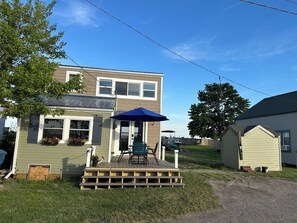 Ein zweistöckiges Haus mit einer Terrasse, einem blauen Sonnenschirm und einem kleinen Gartenhaus.