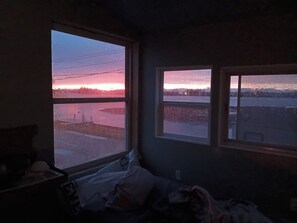 Et værelse med en seng, et vindue med udsigt over en solnedgang hen over vandet, og et andet vindue med udsigt over en by.