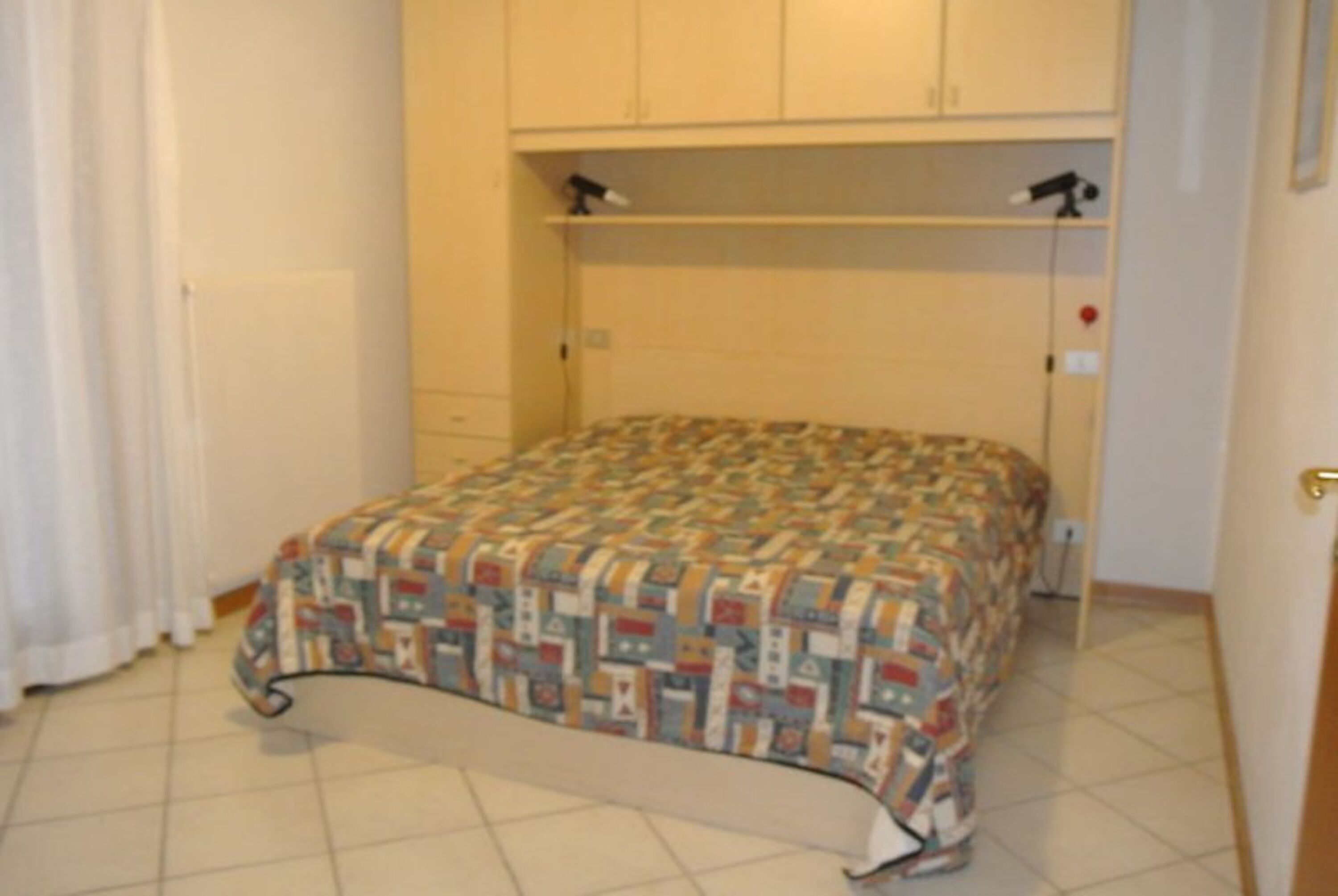 A single bed with a patterned bedspread in a room with a tiled floor and white walls.