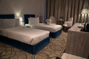 A hotel room with two beds, a desk, a chair, a lamp, and a small table with a vase. - Luxury Palace Hotel (Amman)
