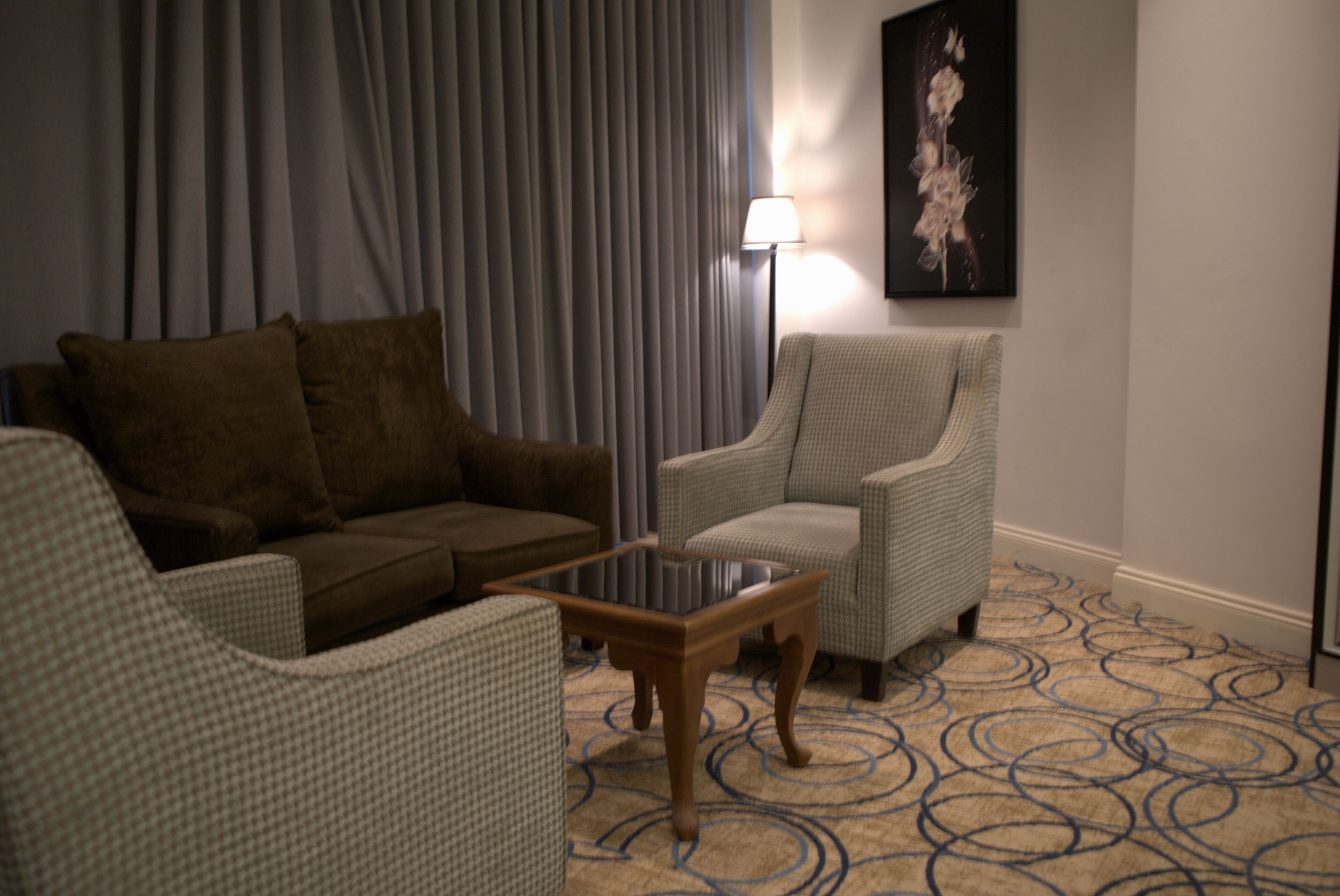 A living room with a brown sofa, a light-colored armchair, a glass-top coffee table, and a patterned carpet.
