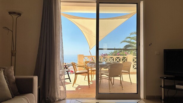A room with a sofa, a TV, and a sliding glass door leading to an outdoor seating area with chairs and a table. - 180 Degree Sea Views, Sunny Terrace & Quiet Pool (Luz)