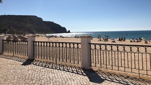 A beach with a paved walkway, a metal fence, and a rocky cliff in the background. - 180 Degree Sea Views, Sunny Terrace & Quiet Pool (Luz)