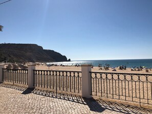Ein Strand mit einer gepflasterten Promenade, einem Metallzaun und einer felsigen Klippe im Hintergrund.
