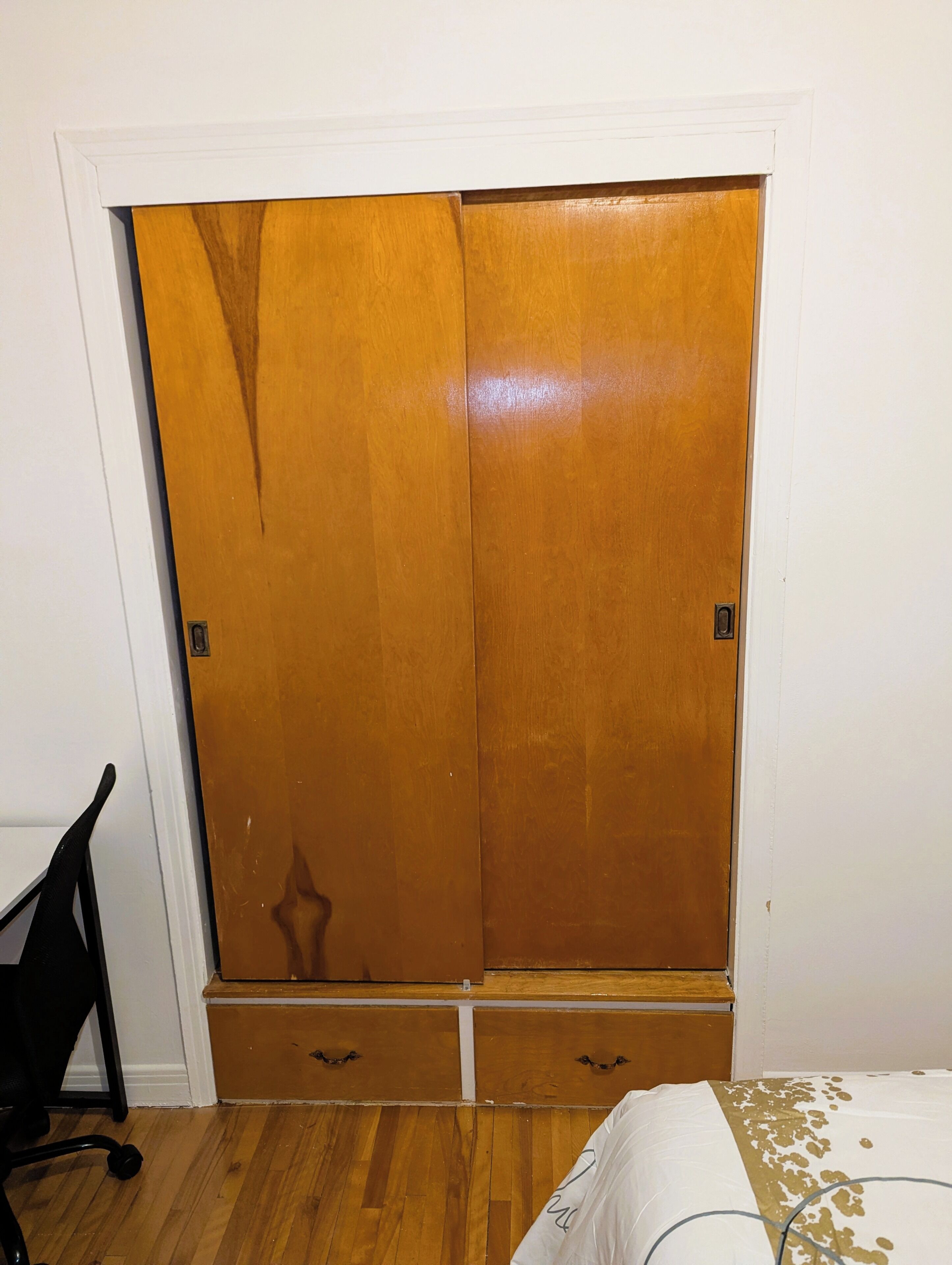 A room with a wooden wardrobe, a black office chair, and a bed with a white and gold bedspread.