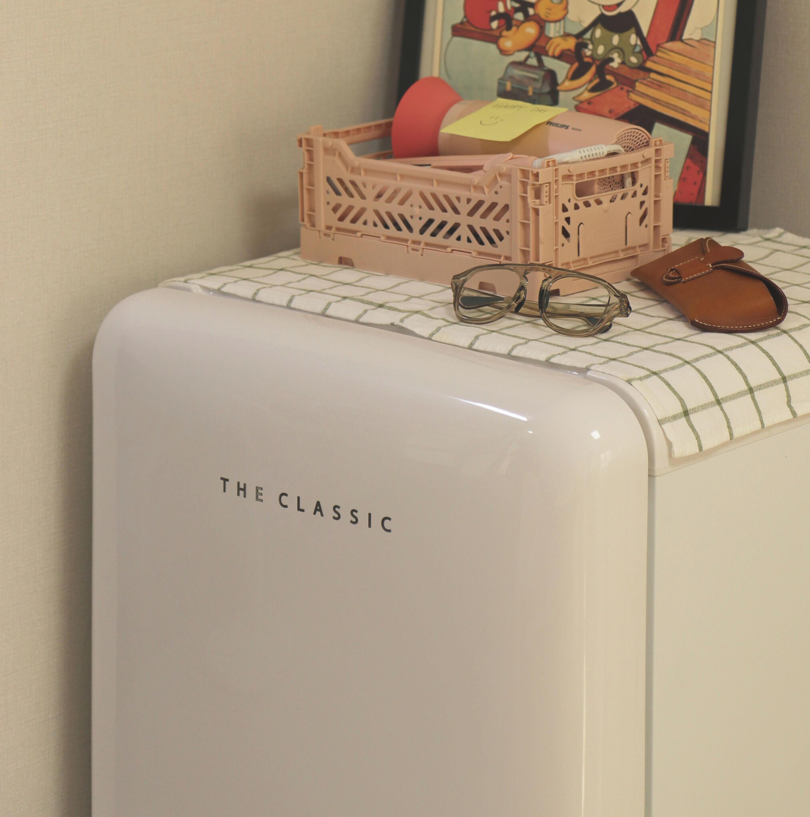 Un electrodoméstico blanco con el texto 'THE CLASSIC', una caja de plástico rosa, un par de gafas y un artículo de cuero marrón sobre una superficie a cuadros.