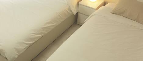 Two single beds with white bedding, a bedside table with a lamp, and a plain wall.