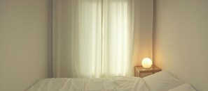 Een bed met wit beddengoed, een nachtkastje met een lamp en dunne gordijnen.