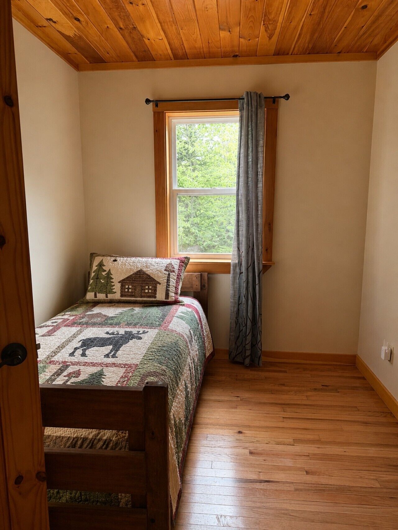 A bedroom with a wooden ceiling, a window with curtains, a bed with a quilt, and a wooden floor.