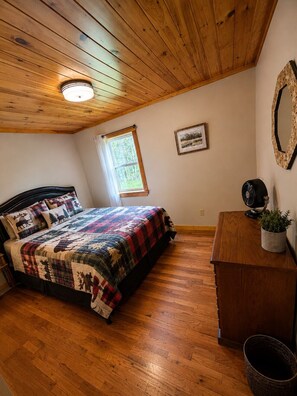 A bedroom with a wooden ceiling, a bed with a quilt, a nightstand, a fan, and a window.