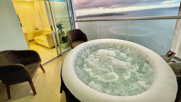 A balcony with a hot tub, wicker chairs, a sofa, and a glass door leading to a room with a yellow wall and a plant. - 1 Bedroom Sunset Luxury Apartment Private Jacuzzi (Cartagena)