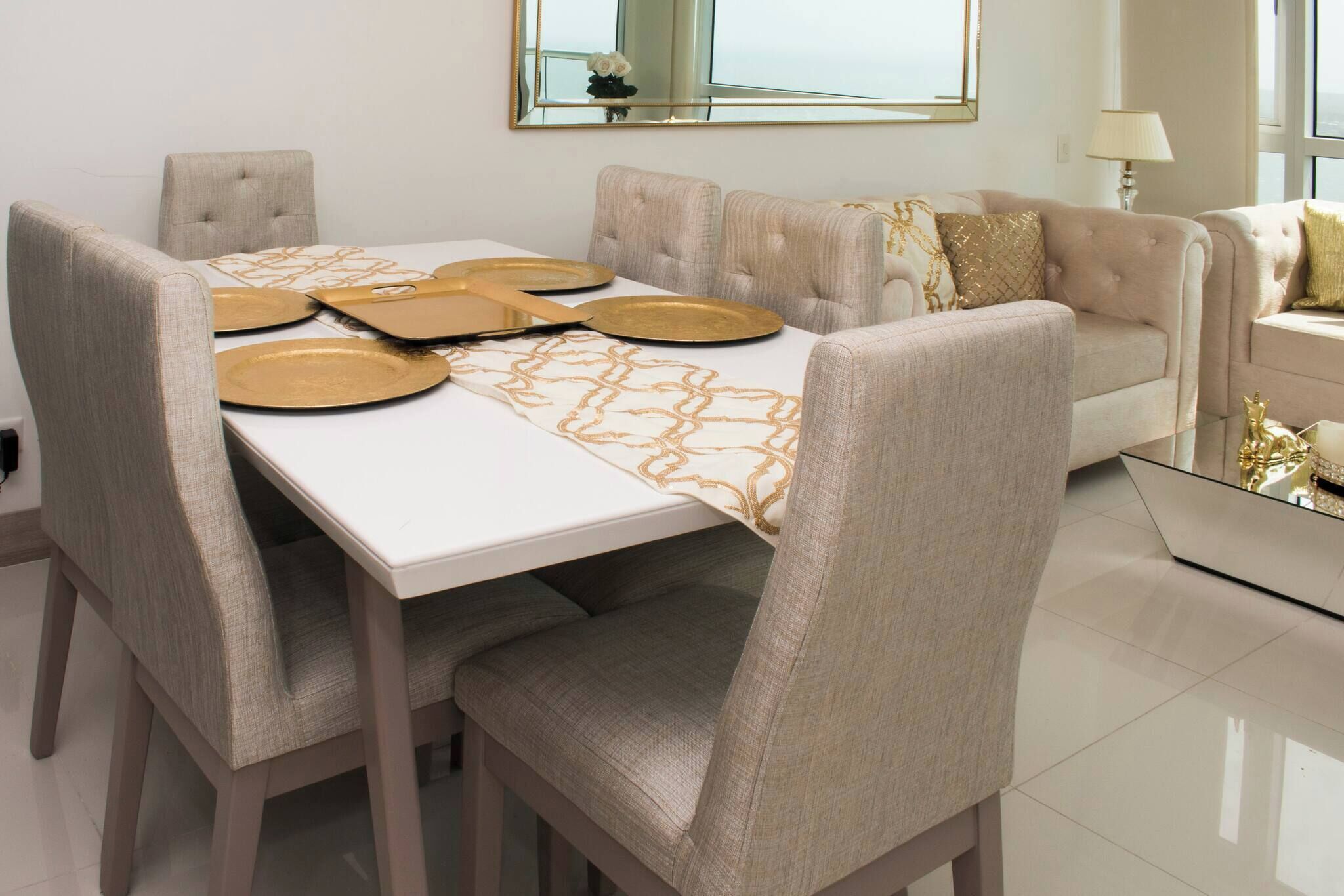 Un área de comedor con una mesa blanca, sillas tapizadas en tono beige y un sofá con cojines estampados.