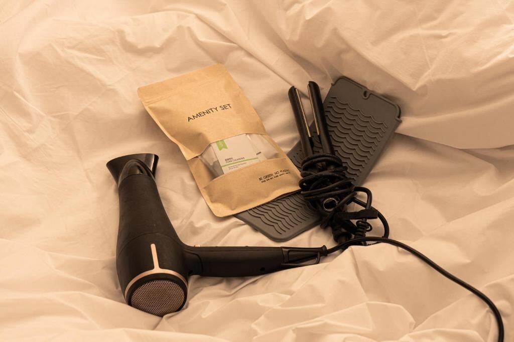 Un secador de pelo, un par de planchas negras y una bolsa de papel marrón con la etiqueta 'AMENITY SET' sobre una superficie de tela blanca.