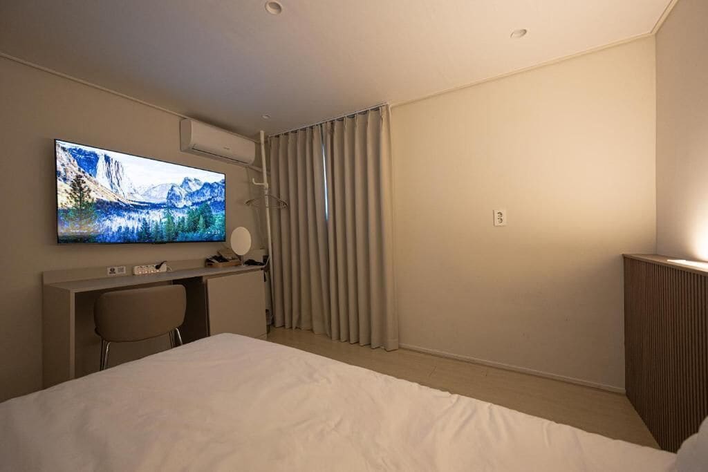 Quarto de hotel com cama, escrivaninha, cadeira, televisão e janela com cortinas.