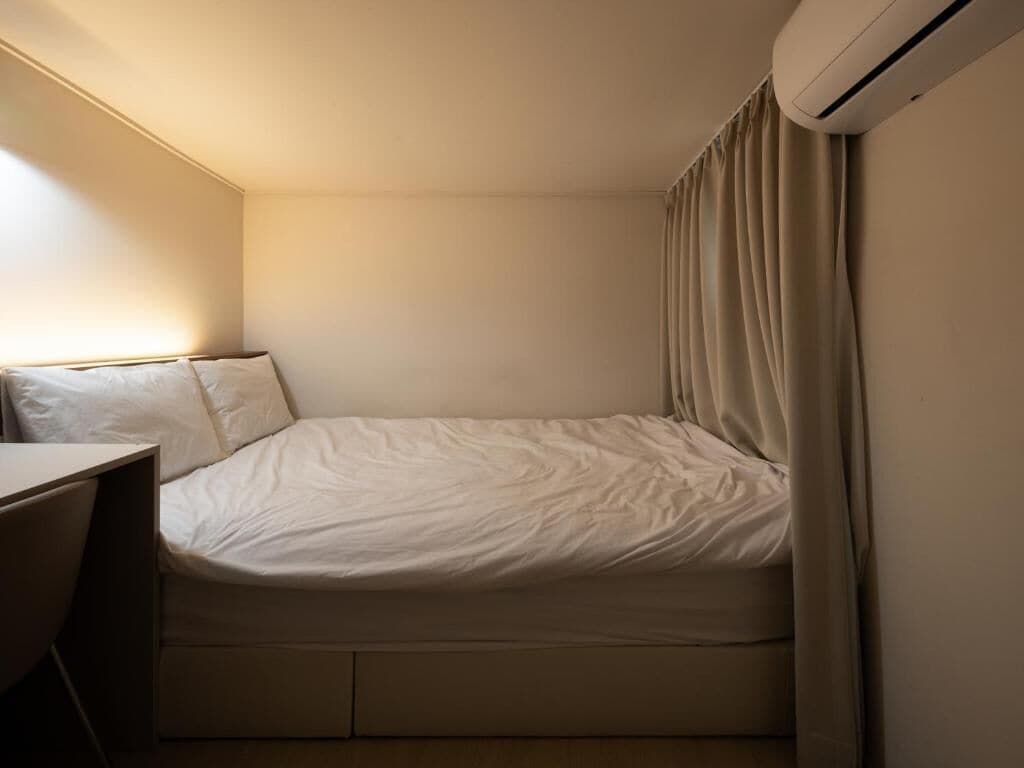 Ein kleines Zimmer mit einem Einzelbett, einem Schreibtisch, einer Wand-Klimaanlage und einem Vorhang.