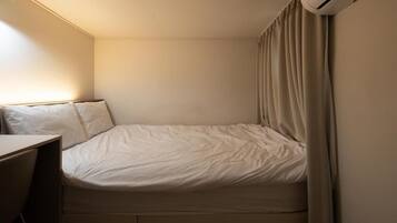 Ein kleines Zimmer mit einem Einzelbett, einem Schreibtisch, einer Wand-Klimaanlage und einem Vorhang.