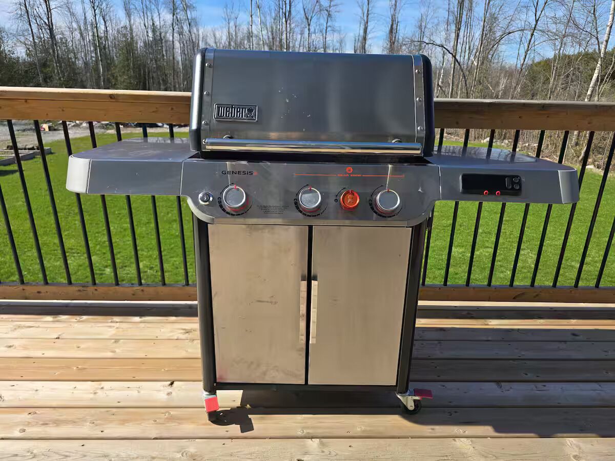 Una parrilla de acero inoxidable para exteriores con pantalla digital, perillas de control y un quemador lateral, ubicada sobre una terraza de madera con barandilla.