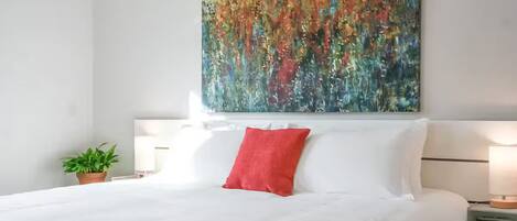 Una cama bien hecha con ropa de cama blanca, una almohada decorativa roja y un cuadro abstracto grande sobre ella.