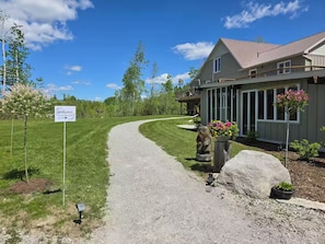 A gravel driveway leading to a house with a porch, a welcome sign, and a well-maintained lawn.