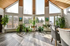 A sunlit room with large windows, wooden flooring, and multiple potted plants.