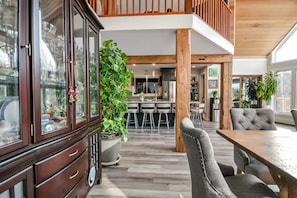 A spacious interior with a wooden dining table, grey upholstered chairs, a large potted plant, and a modern kitchen in the background.