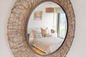 Ein Schlafzimmer mit einem großen runden Spiegel, einem Bett mit vielen Kissen, einer Pendelleuchte und einem Fenster mit Blick auf Bäume.