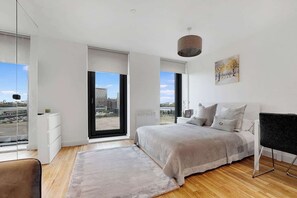Ein modernes Schlafzimmer mit einem großen Bett, einem Schreibtisch und einem Stuhl. Große Fenster bieten Blick auf Gebäude und die Skyline der Stadt.