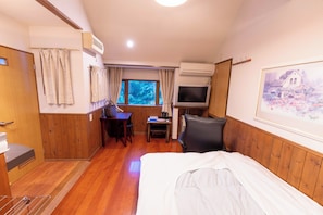 A hotel room with a bed, a desk, a chair, an air conditioning unit, a television, and a window with a view of trees.