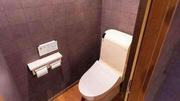 Une petite salle de bain moderne, équipée de toilettes, d’un porte-rouleau de papier toilette et d’une paire de chaussures posées sur le sol.