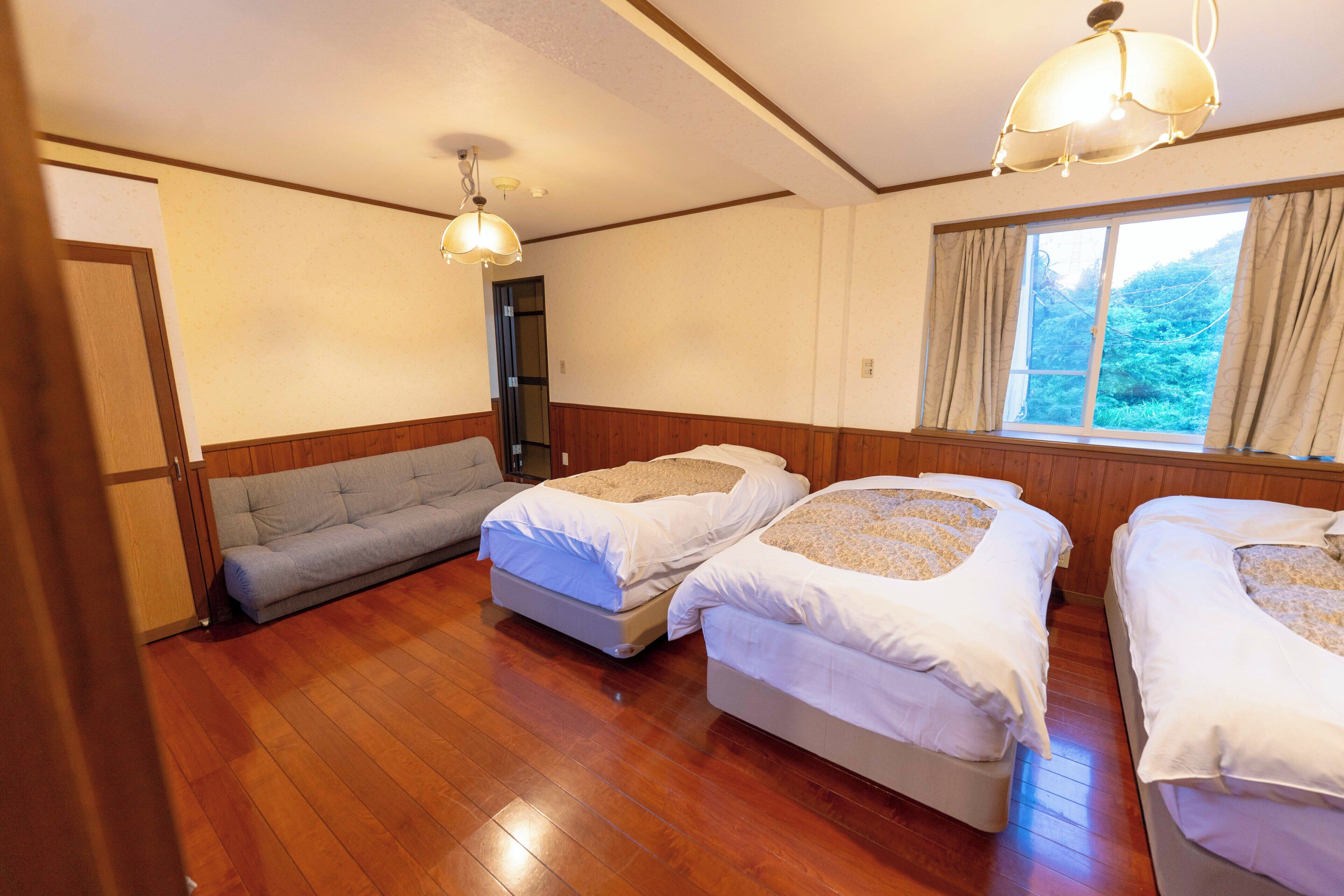 A room with two beds, a sofa, wooden flooring, and a window with curtains.