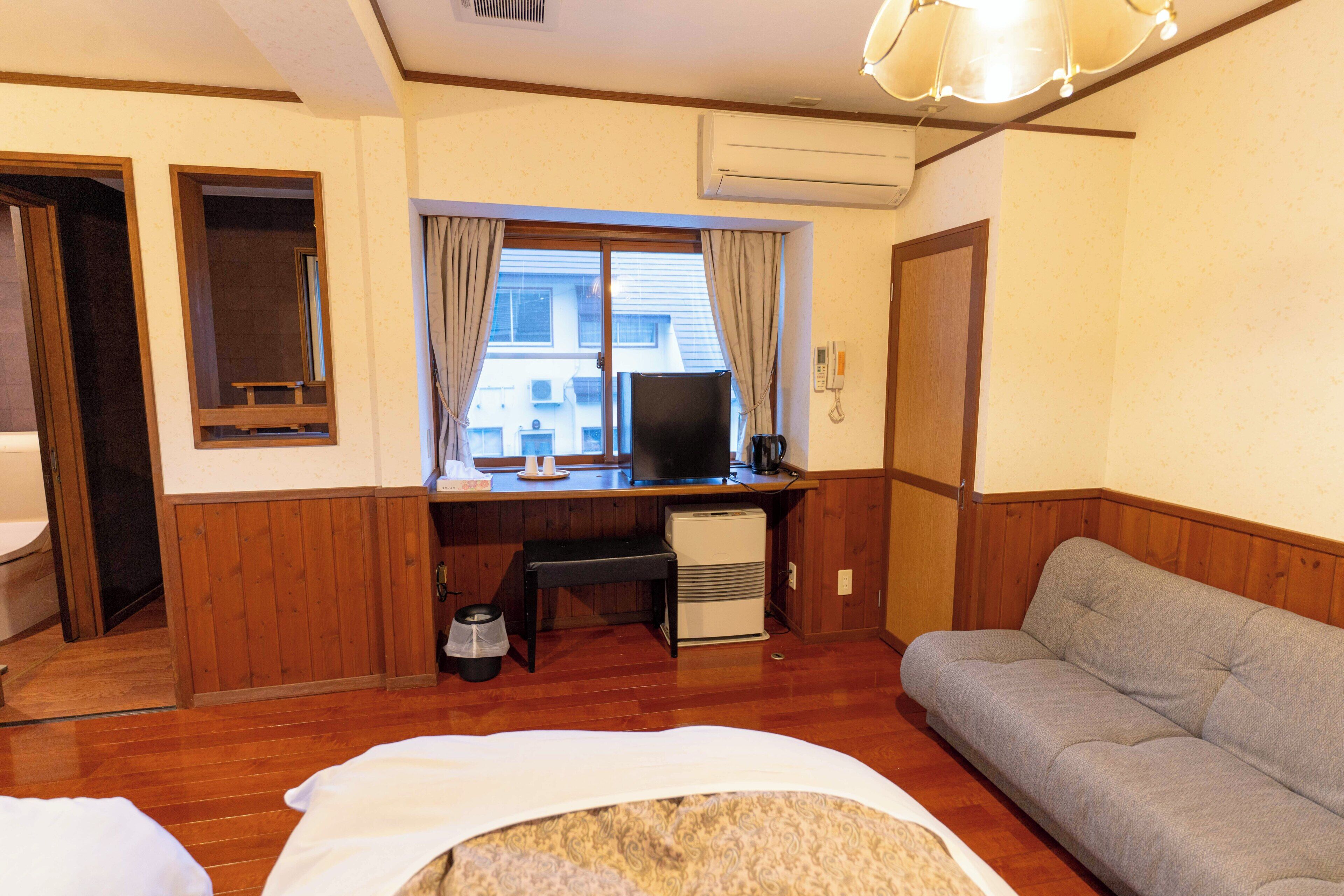 Habitación de hotel con suelo de madera, un sofá, un escritorio con computadora, un calentador pequeño y una ventana con cortinas.