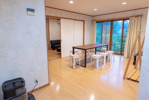 Una habitación con una mesa de madera y cuatro sillas blancas, un sofá negro, y un divisor corredizo que da a otra habitación.