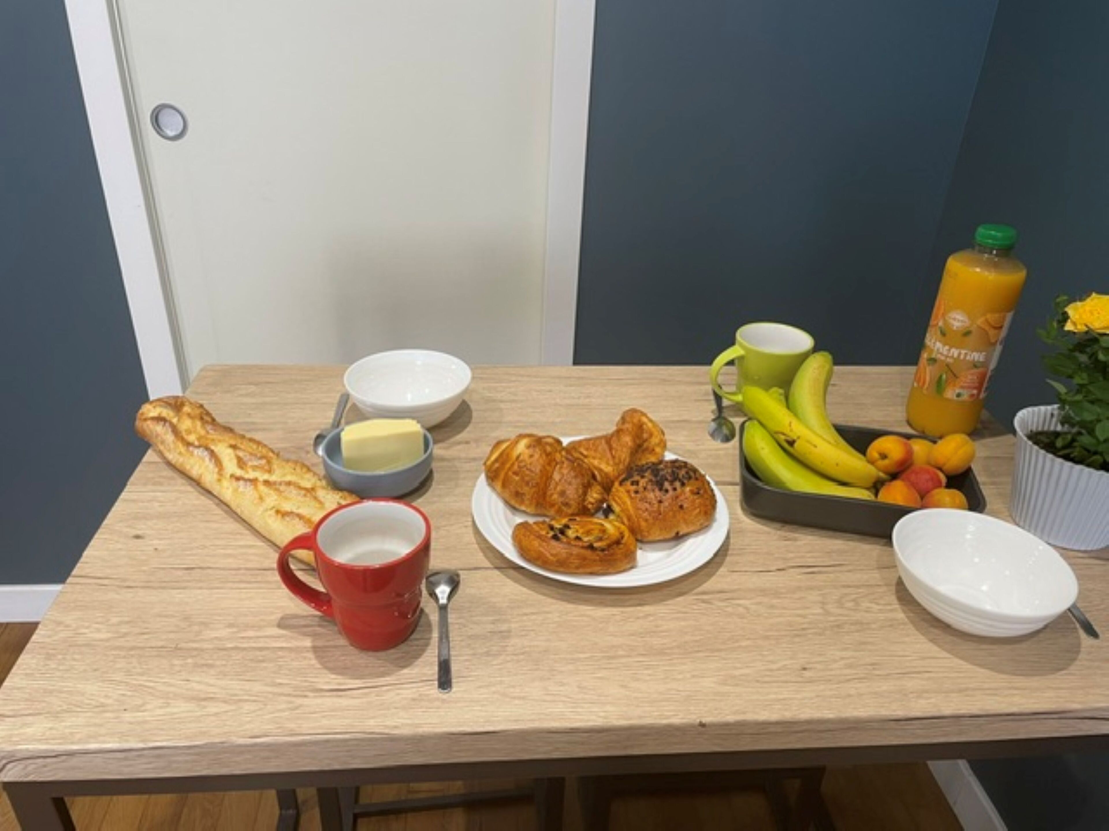 A breakfast table with croissants, bread, a red mug, a plate of pastries, a bowl of butter, a fruit tray, a green mug, and a bottle of orange juice.
