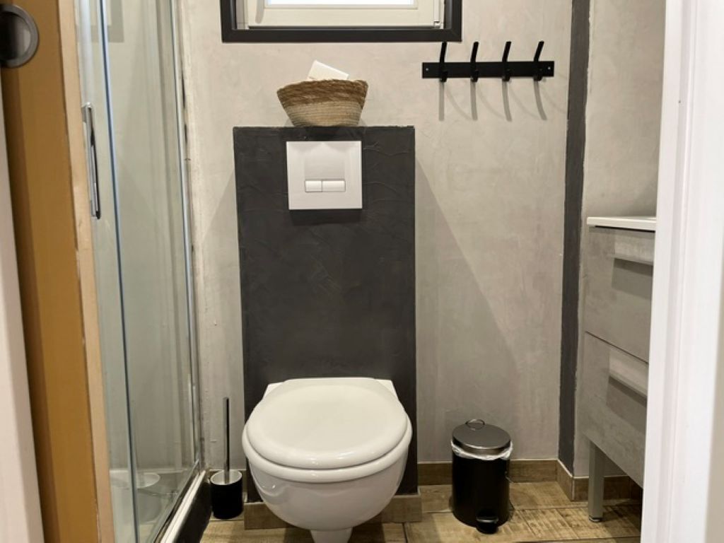 Et badeværelse med toilet, skraldespand, vægmonteret lyskontakt og en krog til at hænge ting på.