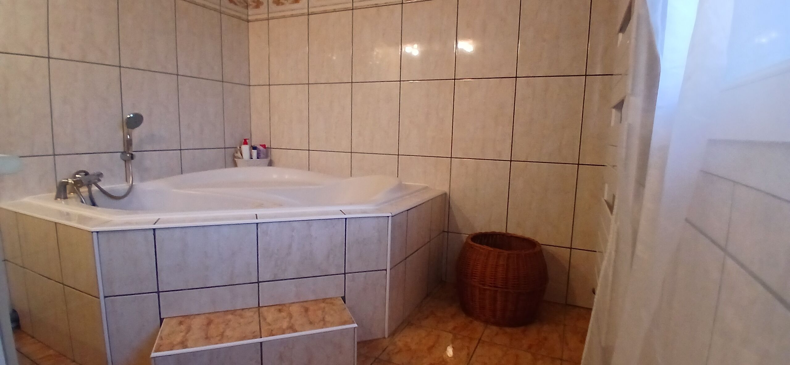 Een badkamer met een badkuip, betegelde muren, een rieten mand en een klein plankje.