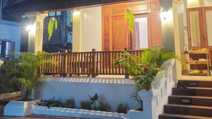 A building with a wooden balcony, steps leading to the entrance, and a view of a hill with a lighted structure in the background. - Loaviengluabgprabang guesthouse (Luang Prabang)